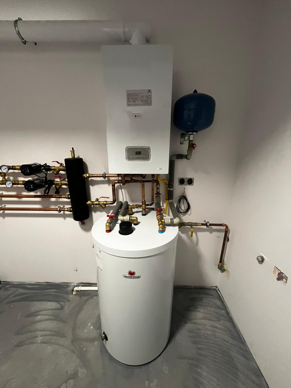 Biały, wiszący piec gazowy Saunier Duval z niebieskim zbiornikiem wyrównawczym i podgrzewaczem wody, widoczne miedziane rury instalacji w surowym pomieszczeniu z betonową podłogą.
