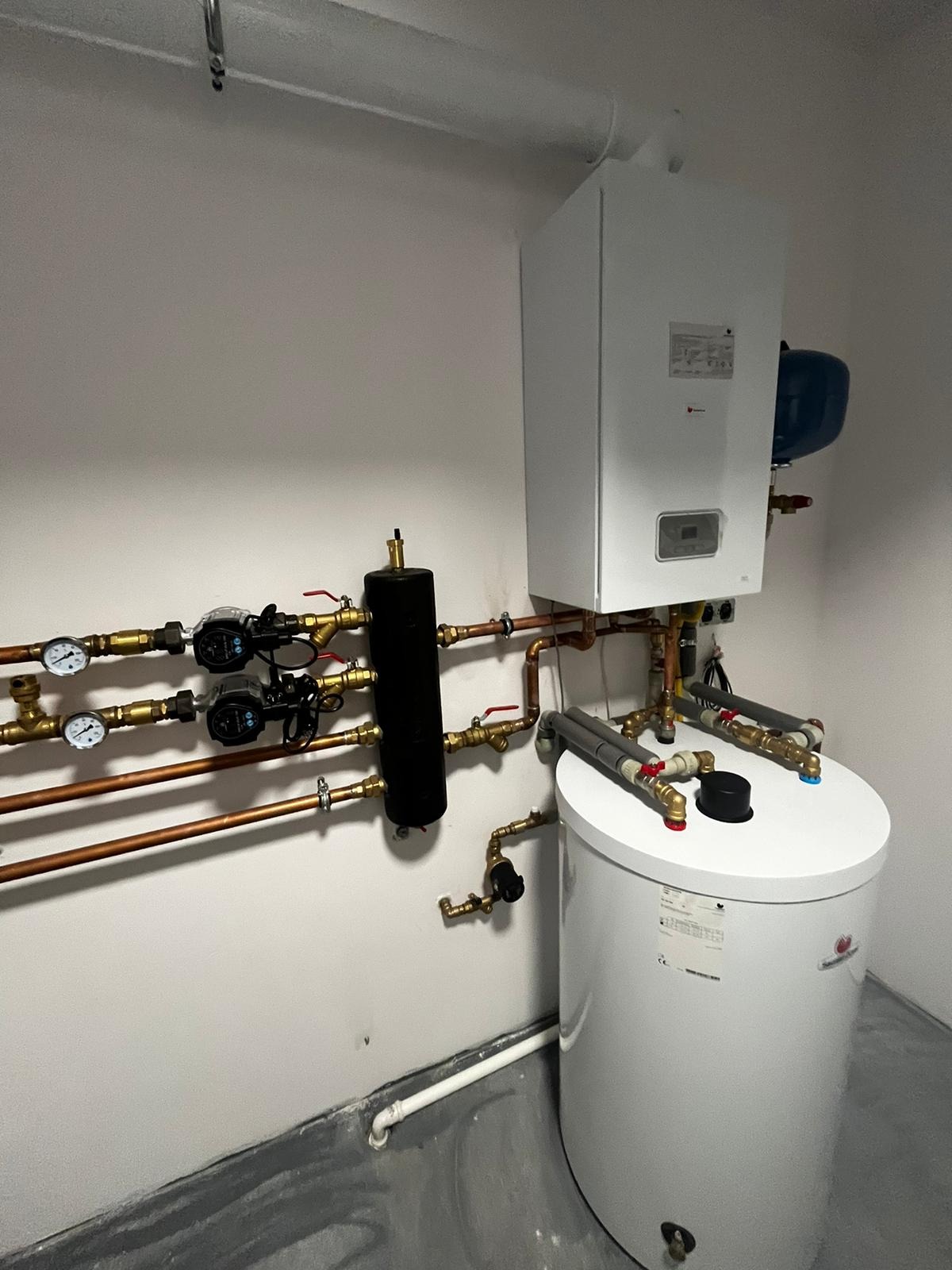 Biały piec gazowy kondensacyjny z podłączonym zasobnikiem ciepłej wody i rozbudowaną instalacją hydrauliczną z pompami obiegowymi i manometrami, zamontowany na ścianie w pomieszczeniu technicznym.