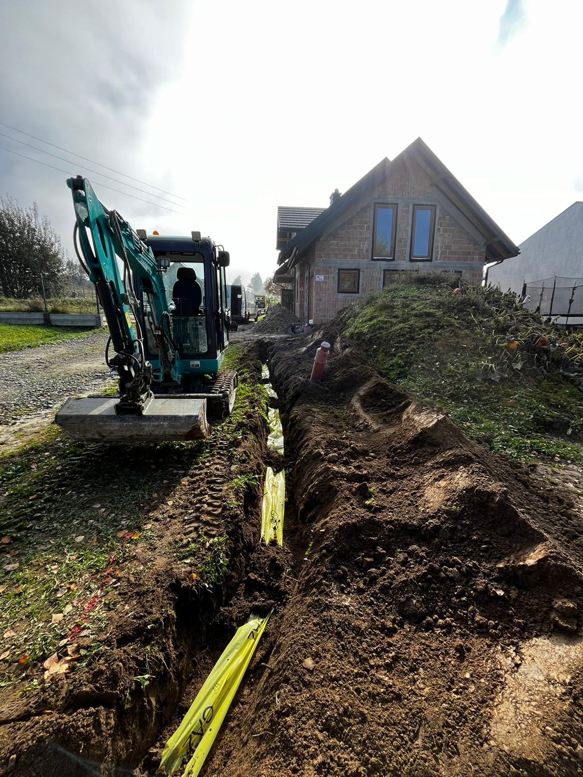 Niebieska mini koparka obok wykopu z żółtą folią ostrzegawczą 'GAZ' w ziemi, widoczny budynek w trakcie budowy w tle.