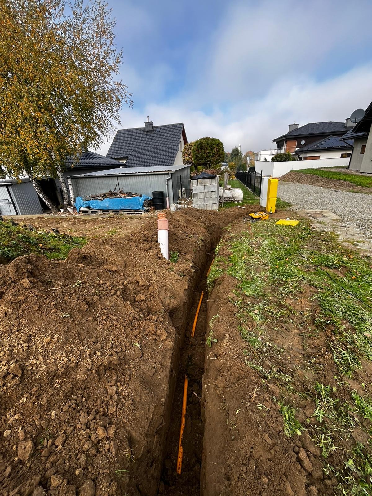 Wykopany rów w ziemi z pomarańczowymi rurami instalacyjnymi na tle zabudowy jednorodzinnej i pochmurnego nieba.