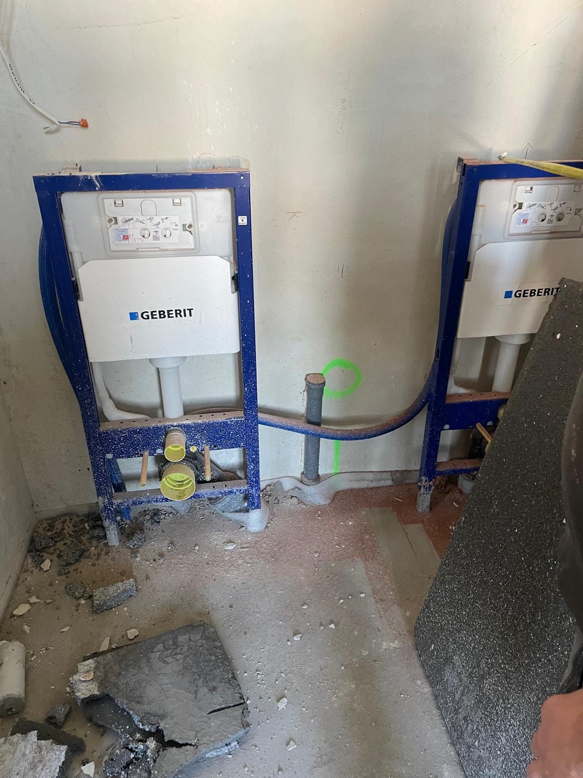 Dwa stelaże podtynkowe Geberit zamontowane w surowym stanie, widoczne rury i przyłącza kanalizacyjne, podłoże z resztkami gruzu i śladami po kuciu.