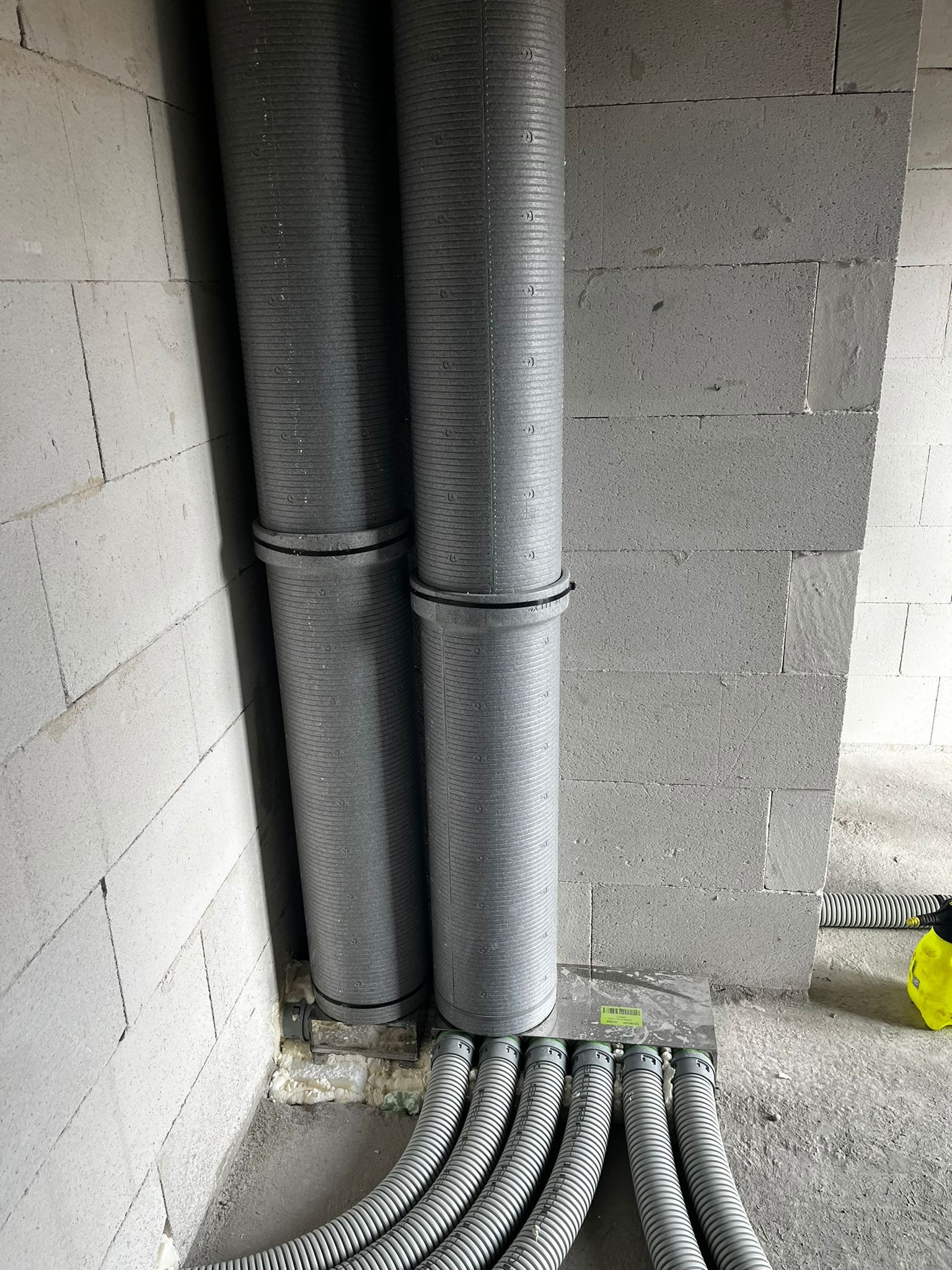 Dwie szare rury kanalizacyjne pionowo przy ścianie z betonu komórkowego, połączone z kilkoma szarymi rurami karbowanymi w podłodze, uszczelnione pianką montażową.