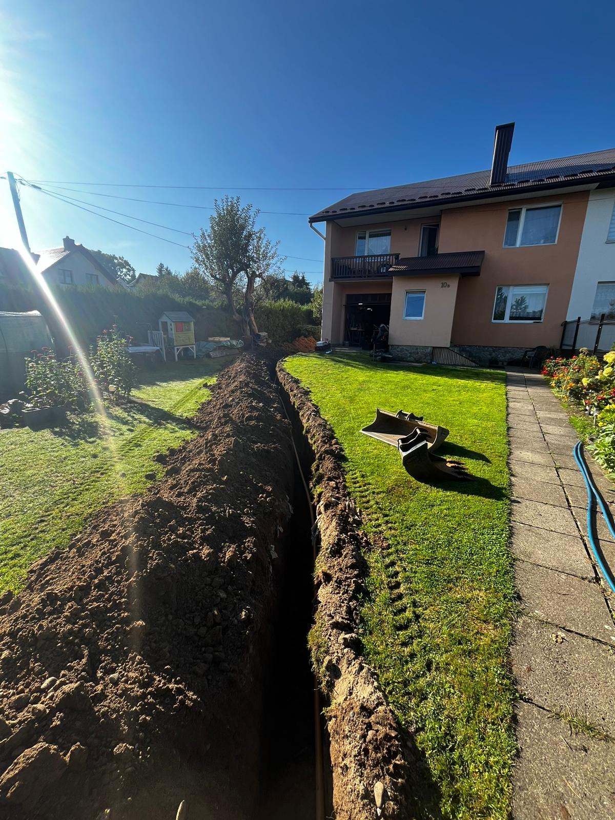 Wykopany rów w trawniku przed domem jednorodzinnym, widoczne dwie łopaty i niebieski wąż, prawdopodobnie przygotowanie pod instalację.