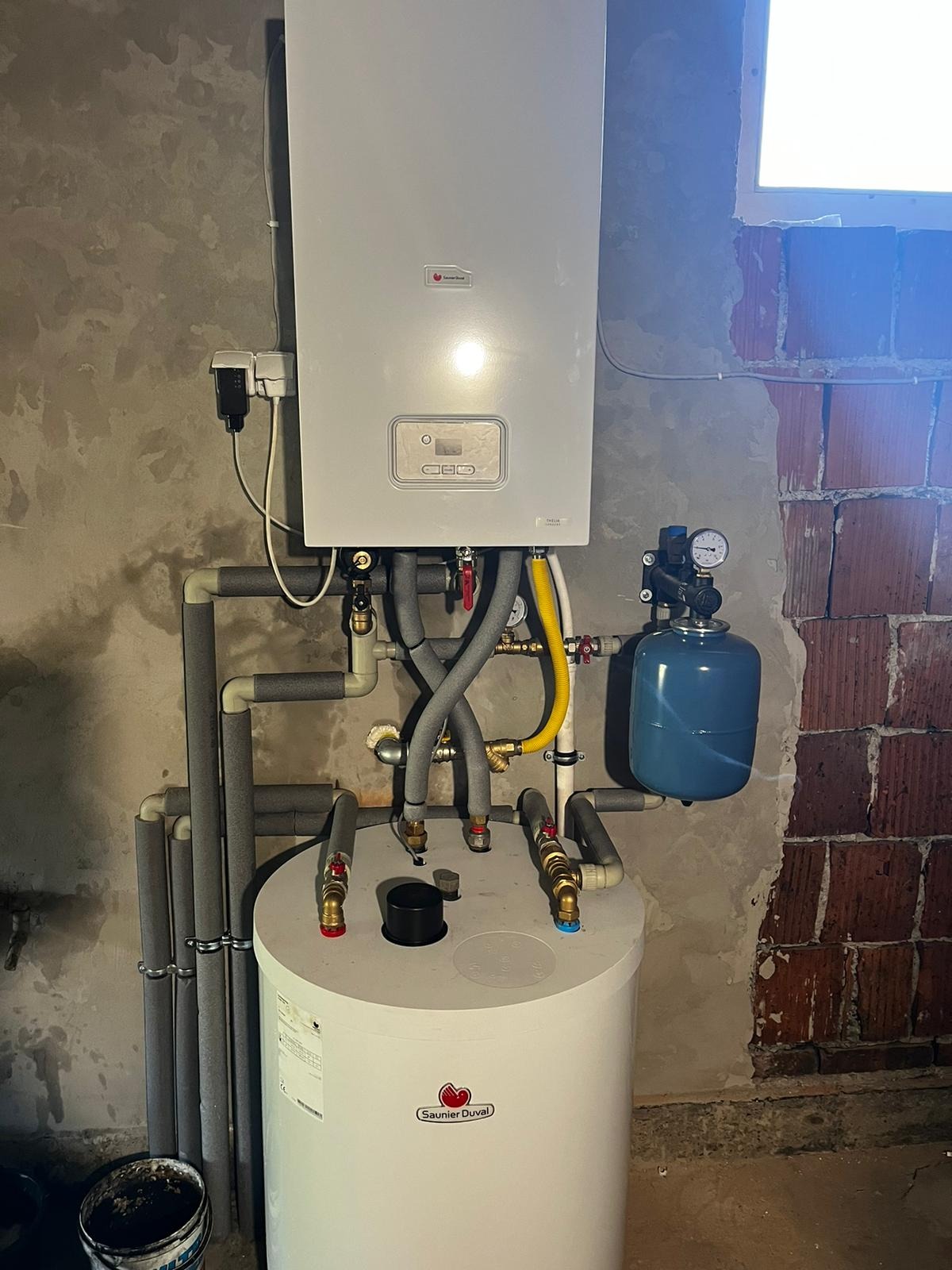 Biały wiszący piec gazowy Saunier Duval z szarymi rurami izolowanymi termicznie, żółtym przewodem gazowym i niebieskim zbiornikiem wyrównawczym na tle surowej ściany z cegły i betonu.