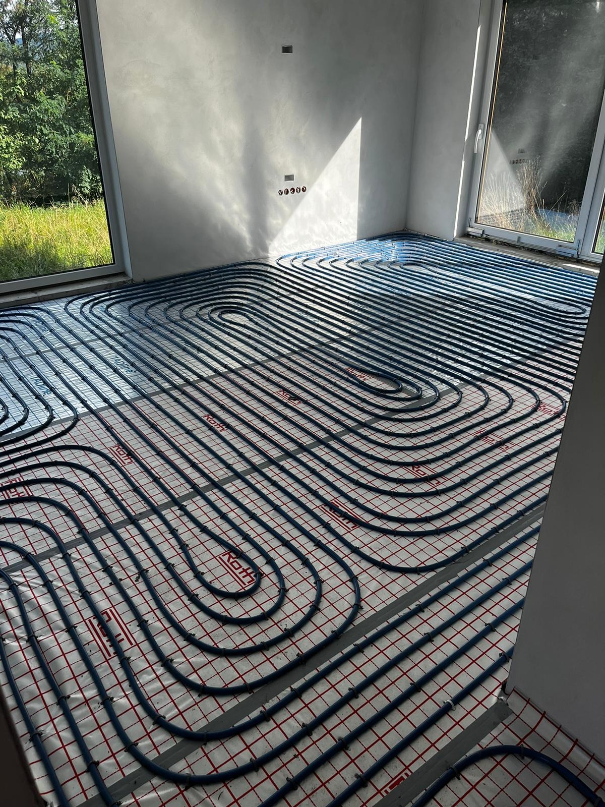 Instalacja ogrzewania podłogowego w nowym budynku, widoczne rury grzewcze ułożone na izolacji z siatką, jasne wnętrze z dużymi oknami.