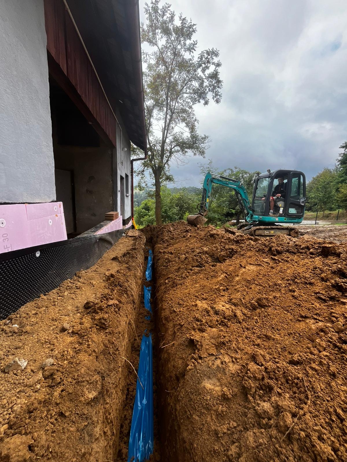 Wykop pod rury, zabezpieczone niebieską folią, wykonany przy budynku z ociepleniem, z użyciem mini koparki.