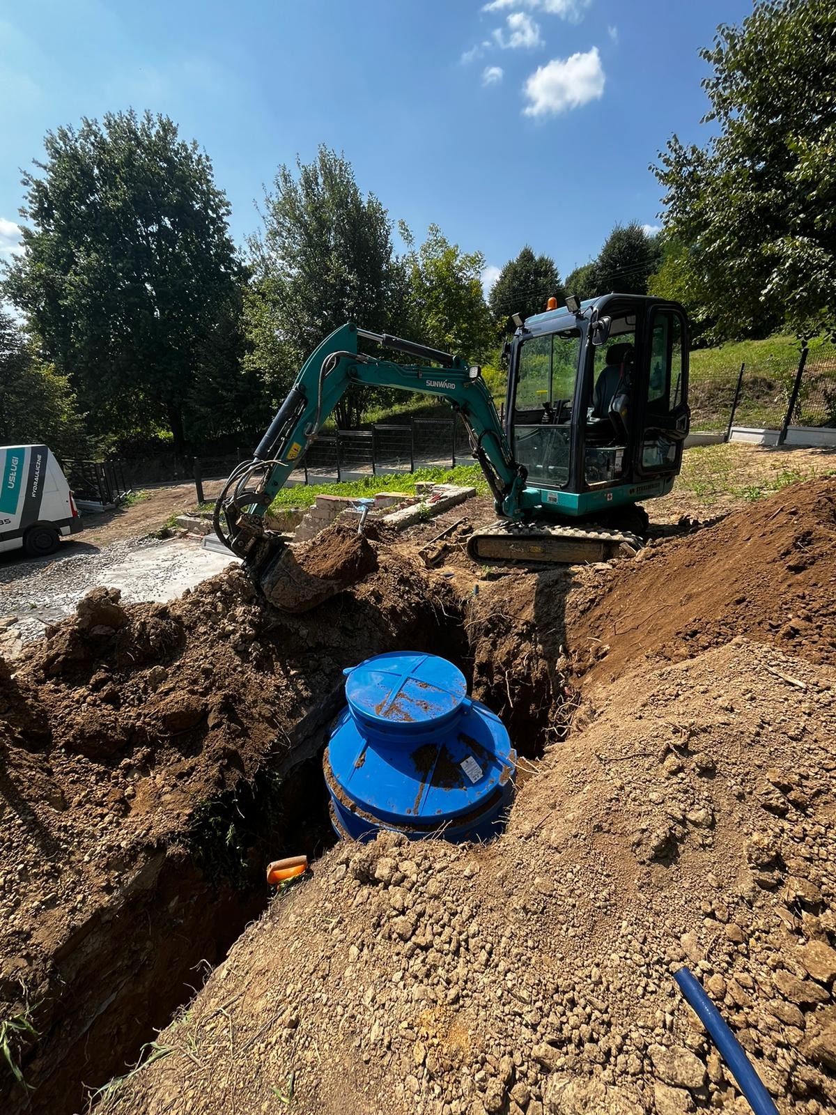 Niebieski zbiornik na ścieki umieszczony w wykopie, obok stoi mała koparka, teren budowy otoczony zielenią.