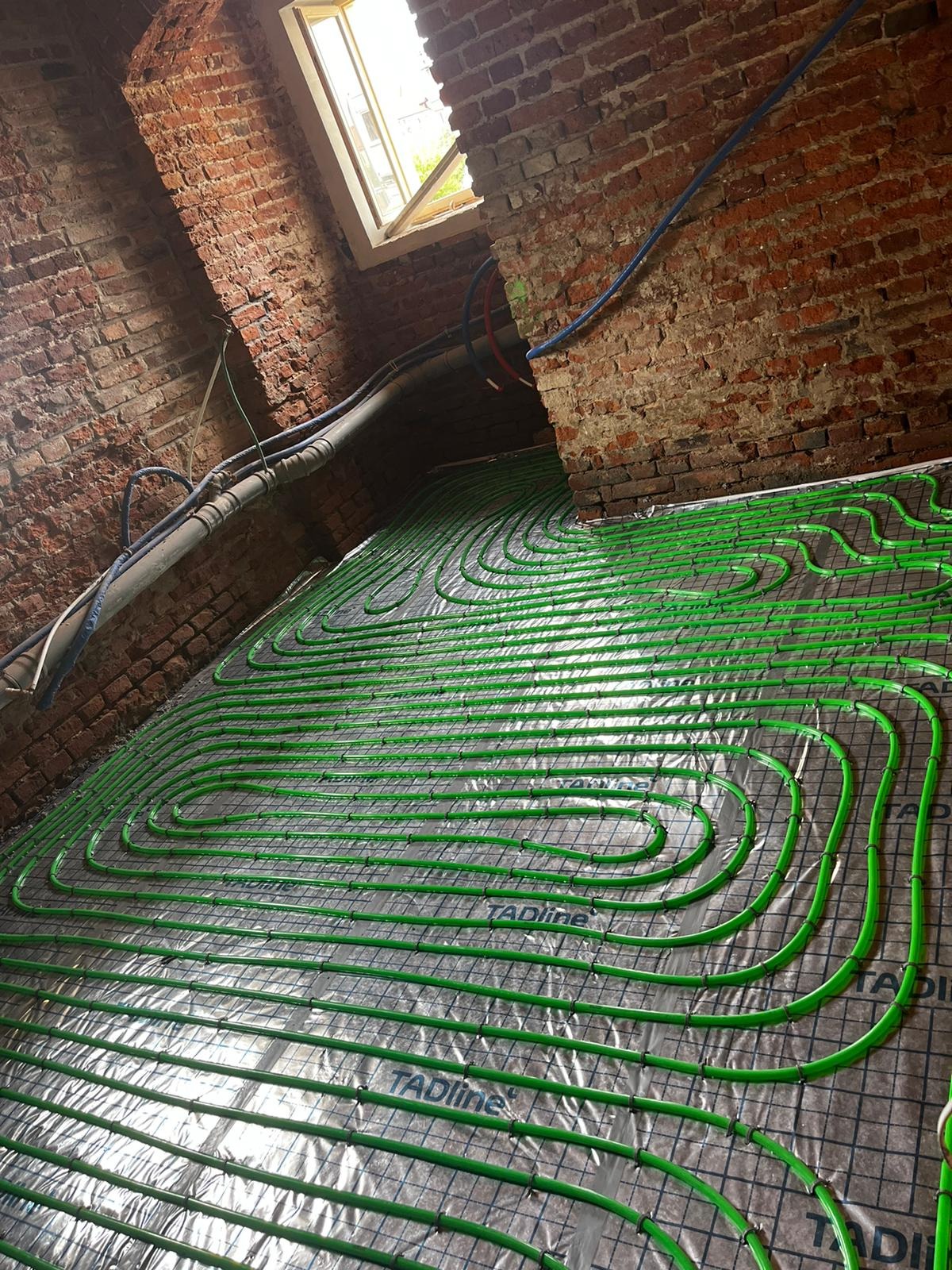Zielone rury ogrzewania podłogowego ułożone spiralnie na folii izolacyjnej z siatką, widoczne fragmenty ceglanej ściany i okno dachowe w surowym wnętrzu.