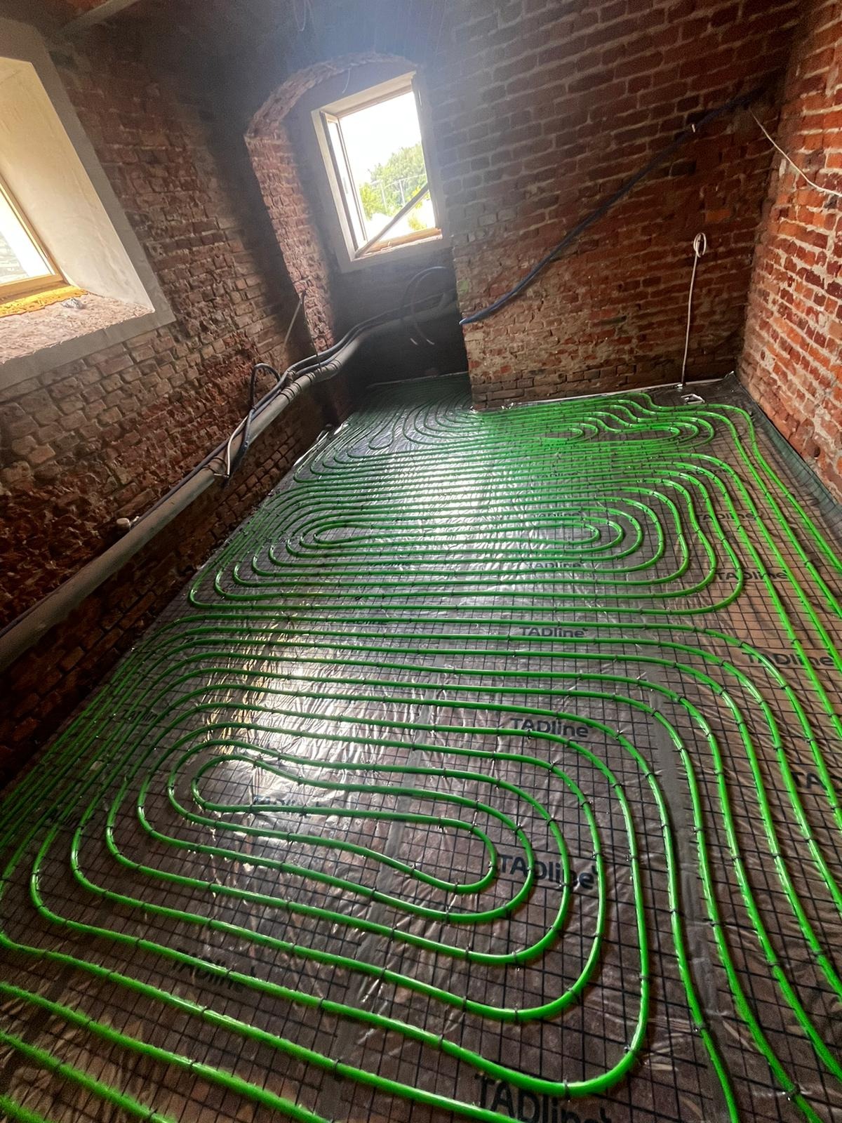 Układanie zielonych rur ogrzewania podłogowego na folii z kratką w pomieszczeniu z ceglanymi ścianami i oknami dachowymi.