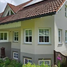 Odnowiona elewacja domu jednorodzinnego z brązową dachówką, białymi oknami i zadbanym ogrodem z ozdobnym żeliwnym elementem przed wejściem.