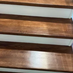 Błyszczące, drewniane schody z białymi podstopnicami, ukazane z bliska pod kątem. Widoczne detale słojów drewna i odbicia światła na powierzchni stopni.