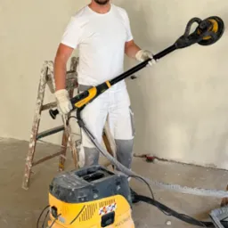 Mężczyzna w bieli szlifuje ścianę szlifierką z odkurzaczem, drabina w tle. Przygotowanie ściany do malowania, etap prac wykończeniowych.