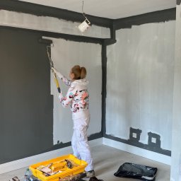 ILLUMINARTI SPÓŁKA Z OGRANICZONĄ ODPOWIEDZIALNOŚCIĄ - Malowanie ścian w pokoju: kobieta maluje ścianę na szaro wałkiem, widać narzędzia malarskie i fragmenty ścian w kolorze białym/szarym.