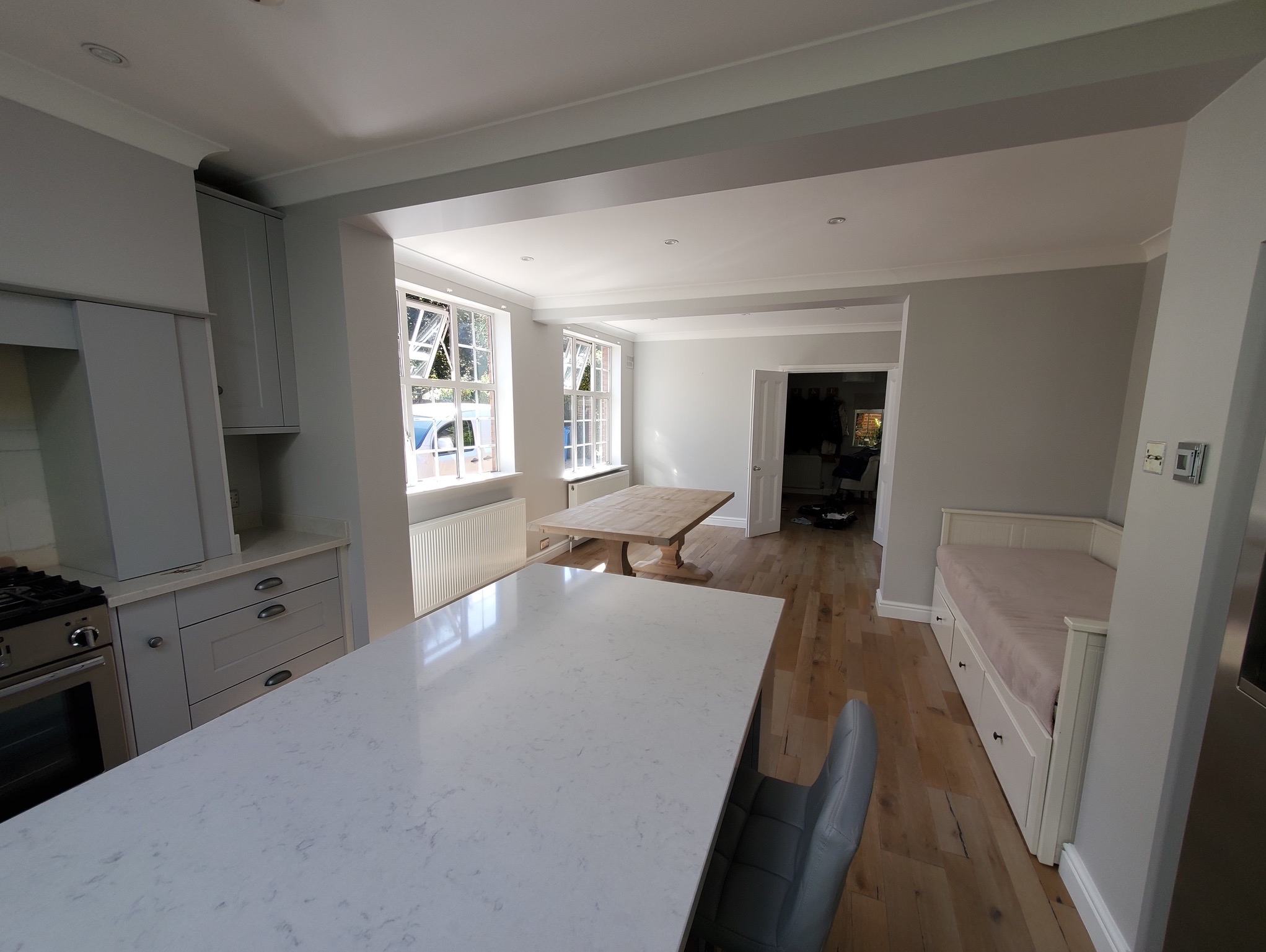 Jasne wnętrze po remoncie: kuchnia z wyspą i salon z drewnianym stołem, ściany pomalowane na szaro, widoczne detale wykończeniowe.