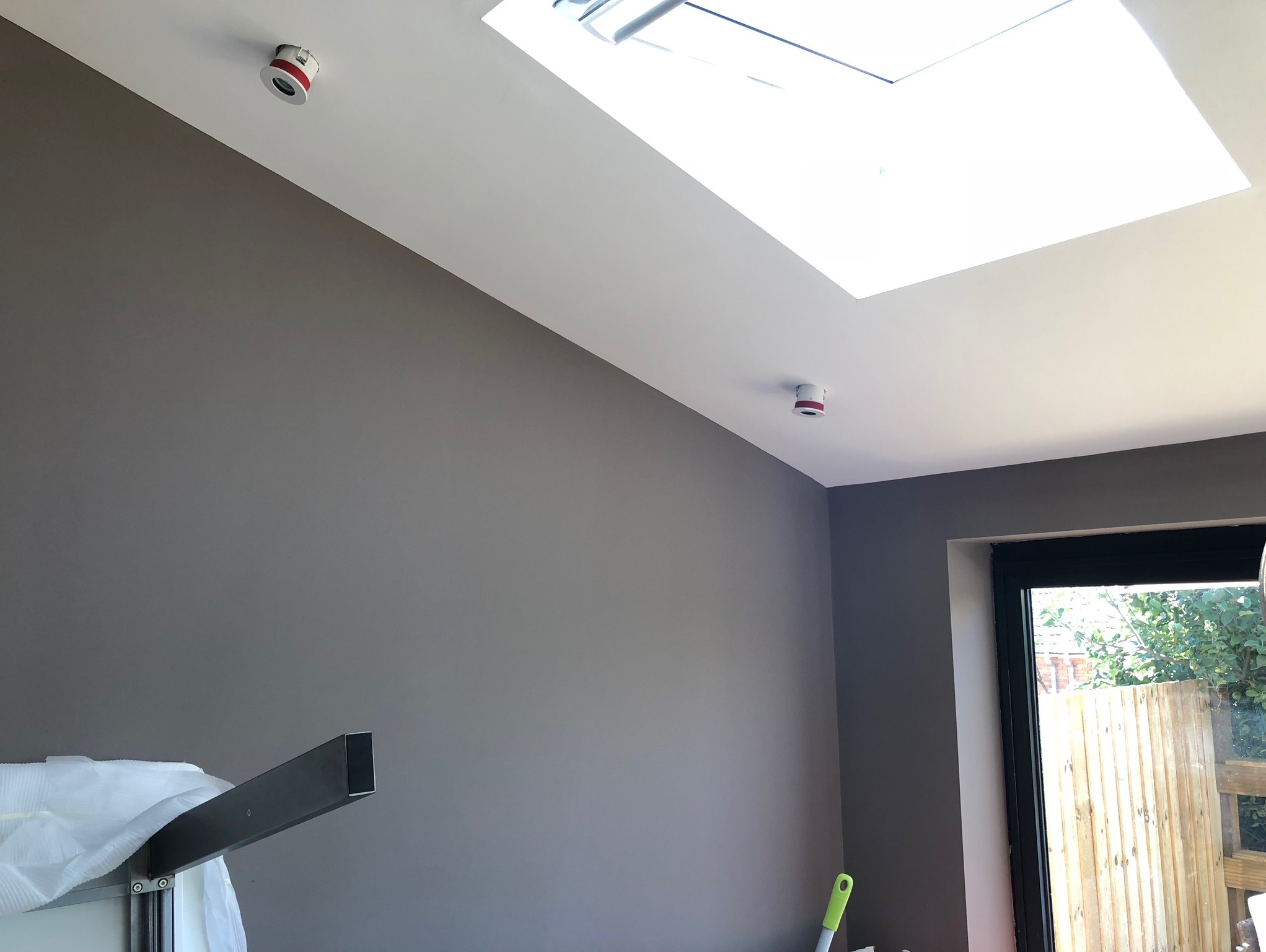 Świeżo pomalowane szare ściany wnętrza z białym sufitem i oknem dachowym; widoczne czujniki dymu na suficie oraz fragment okna i sprzętu w dolnej części kadru.