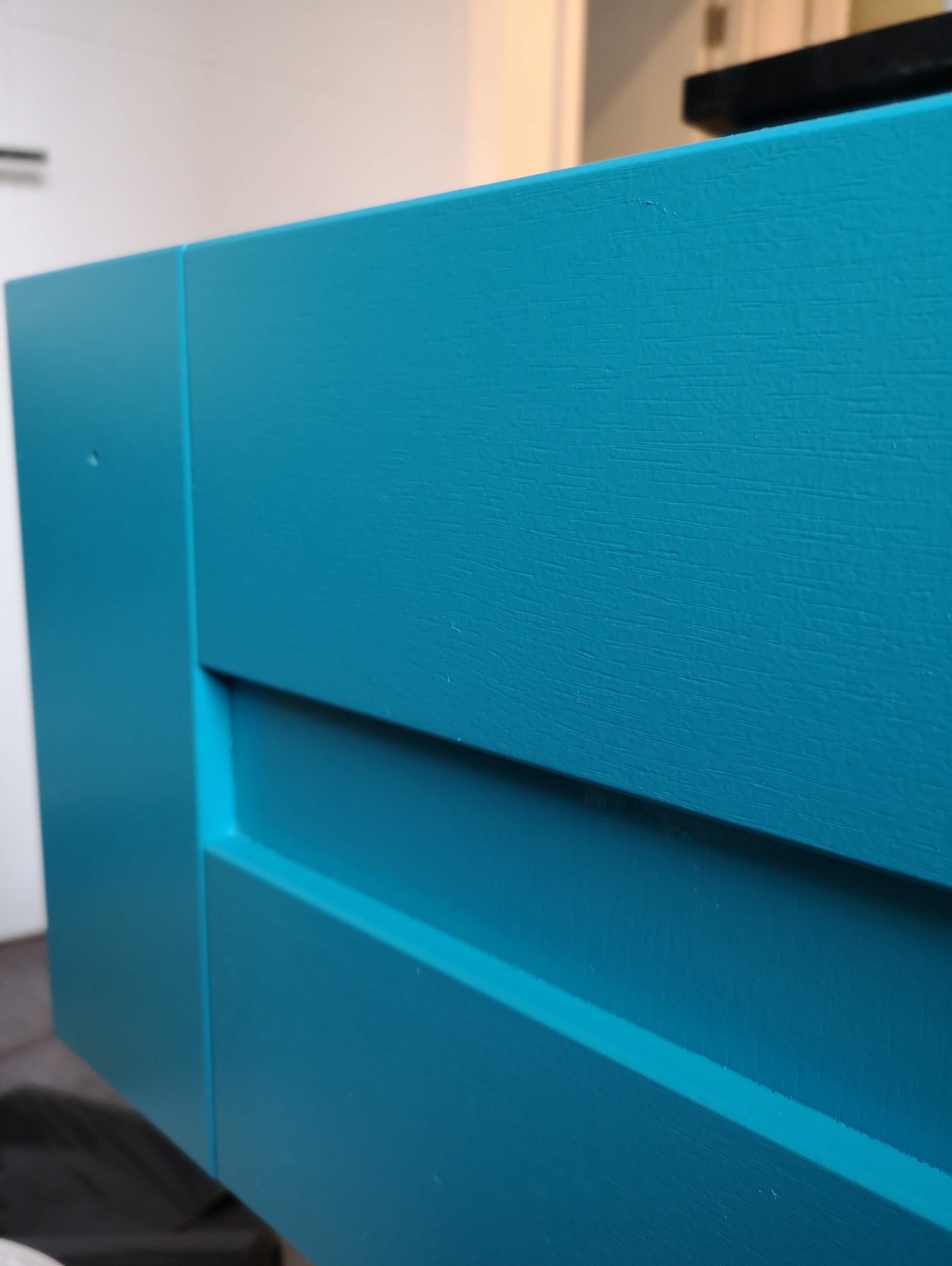 Fragment pomalowanej na turkusowo drewnianej komody z widoczną strukturą drewna i delikatnymi niedoskonałościami powierzchni.