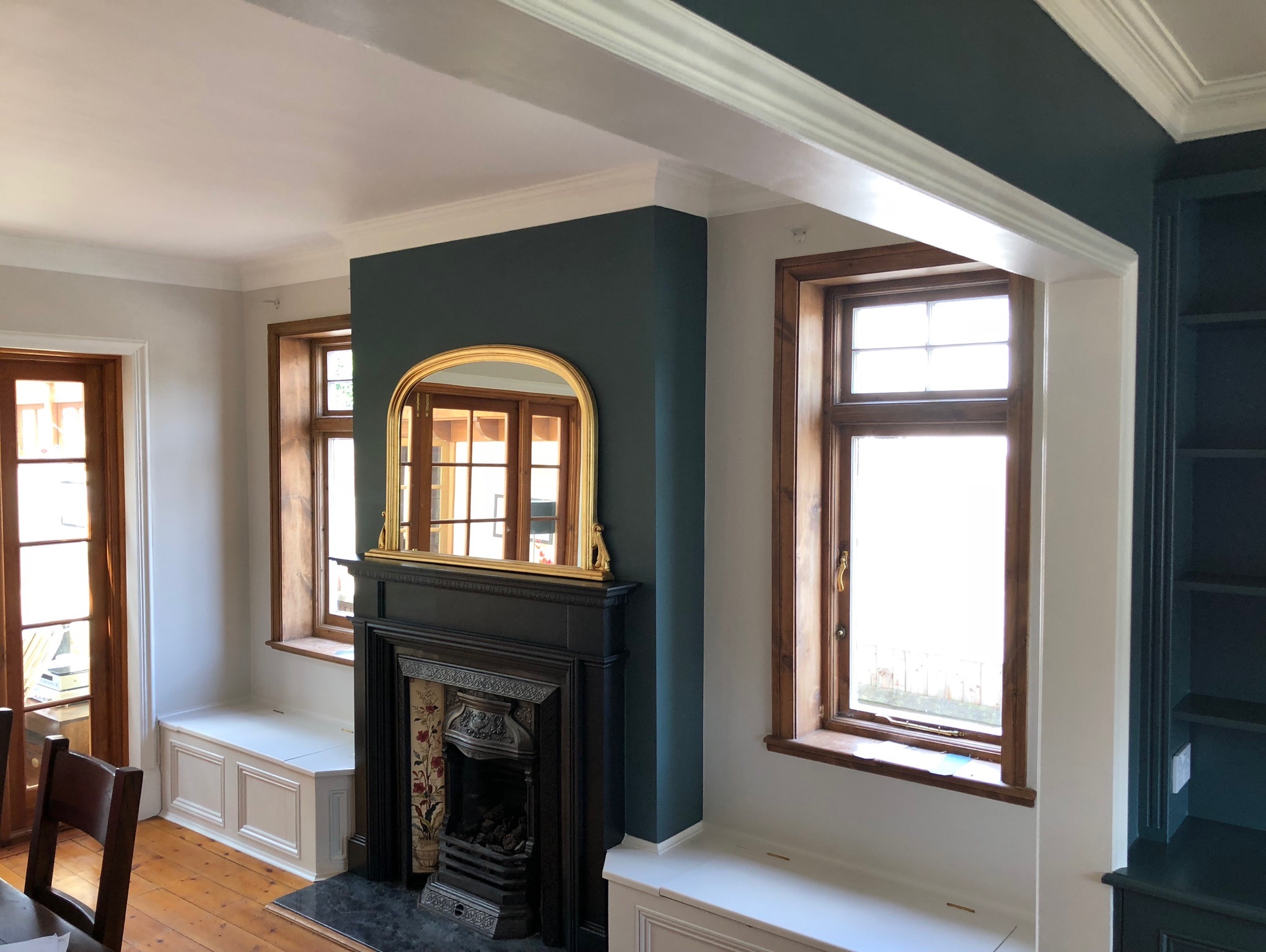 Pomieszczenie po remoncie: ciemnozielona ściana z kominkiem i złotym lustrem, drewniane okna i drzwi, białe listwy przypodłogowe i obudowy okien, drewniana podłoga i zabudowane siedziska pod oknami.