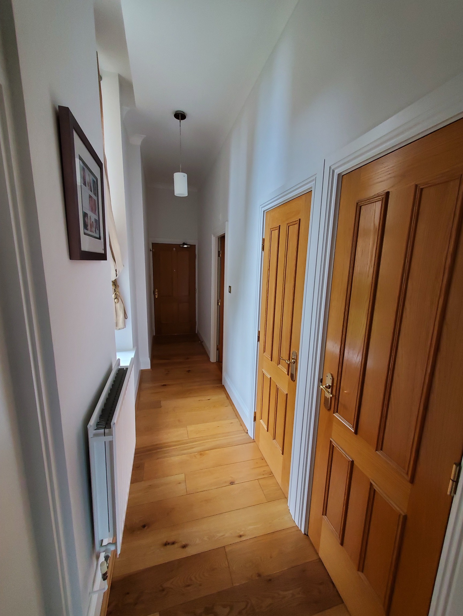 Korytarz po remoncie: świeżo pomalowane białe ściany, lakierowana drewniana podłoga, dwoje drewnianych drzwi z białymi framugami, biały grzejnik i wisząca lampa cylindryczna.