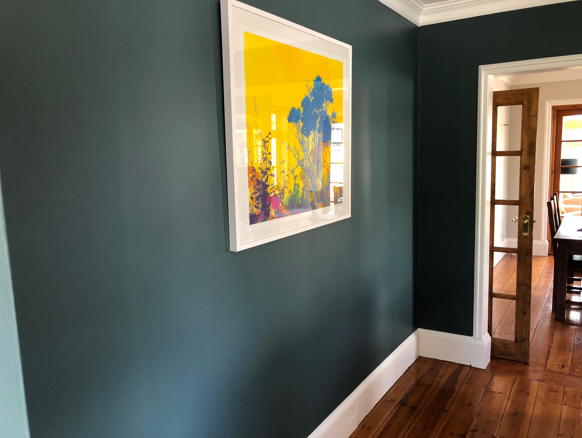 Ściana pomalowana na ciemnozielony kolor z białym obramowaniem u góry i dołu, na ścianie wisi oprawiony obraz z abstrakcyjnym motywem roślinnym w odcieniach żółci i niebieskiego, w tle widoczne...