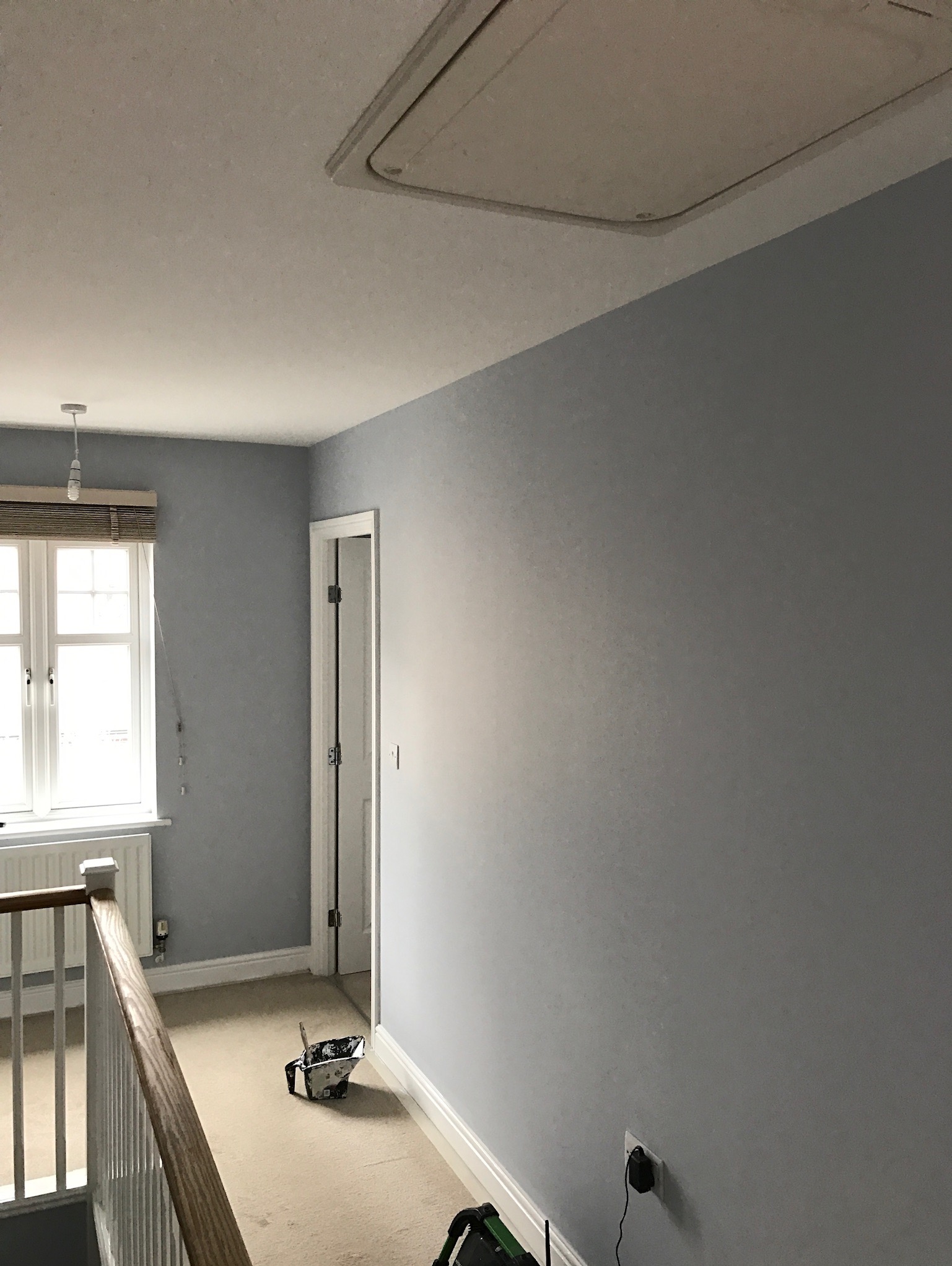 Świeżo pomalowane na szaro wnętrze domu z widocznym oknem z żaluzjami, drzwiami i otwartym włazem na poddasze, z akcesoriami malarskimi leżącymi na dywanie.