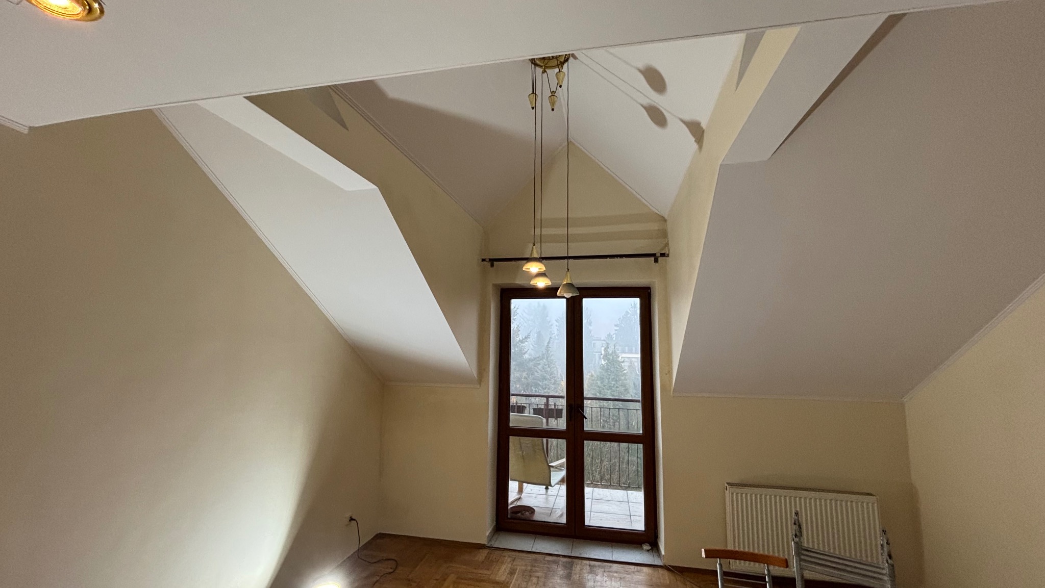 Poddasze z oknem balkonowym, ścianami w odcieniach beżu i skośnym sufitem, widoczny fragment parkietu, wisząca lampa z trzema kloszami i grzejnik.