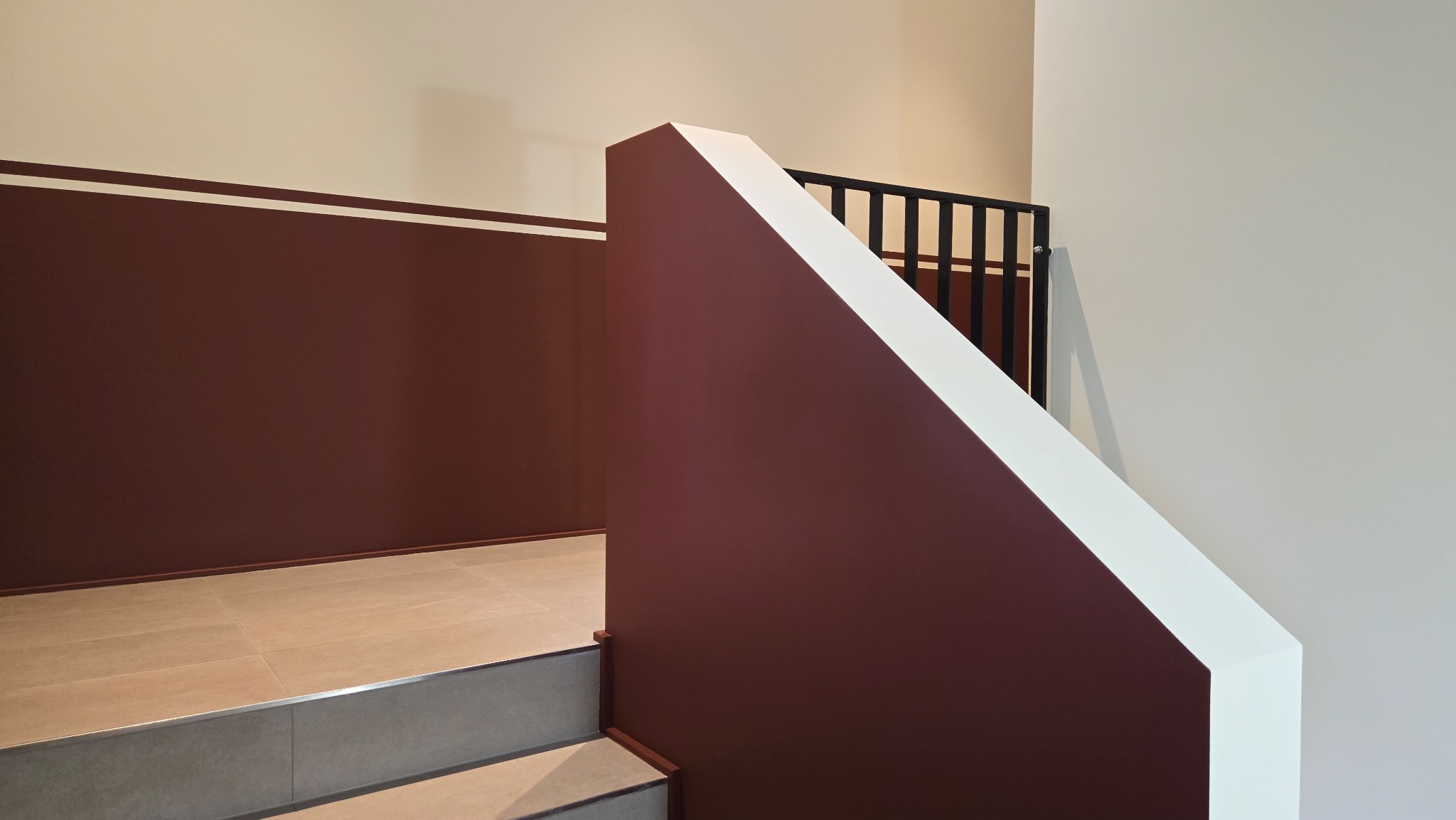Klatka schodowa z bordową ścianą z białym paskiem, jasnymi płytkami na podłodze i czarną balustradą.