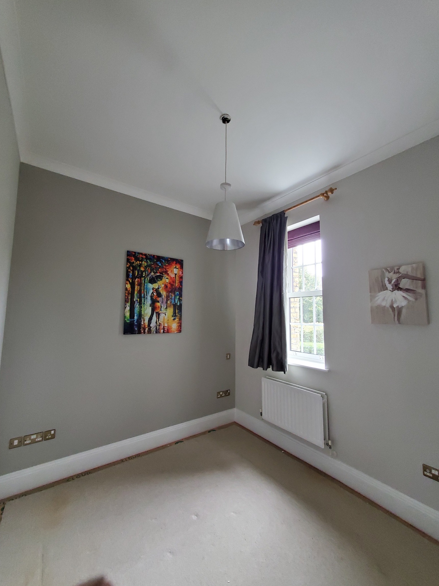 Pomalowane na szaro wnętrze pokoju z białym sufitem i listwami, wisząca lampa z szarym kloszem, obrazy na ścianach, grzejnik pod oknem z zasłonami.