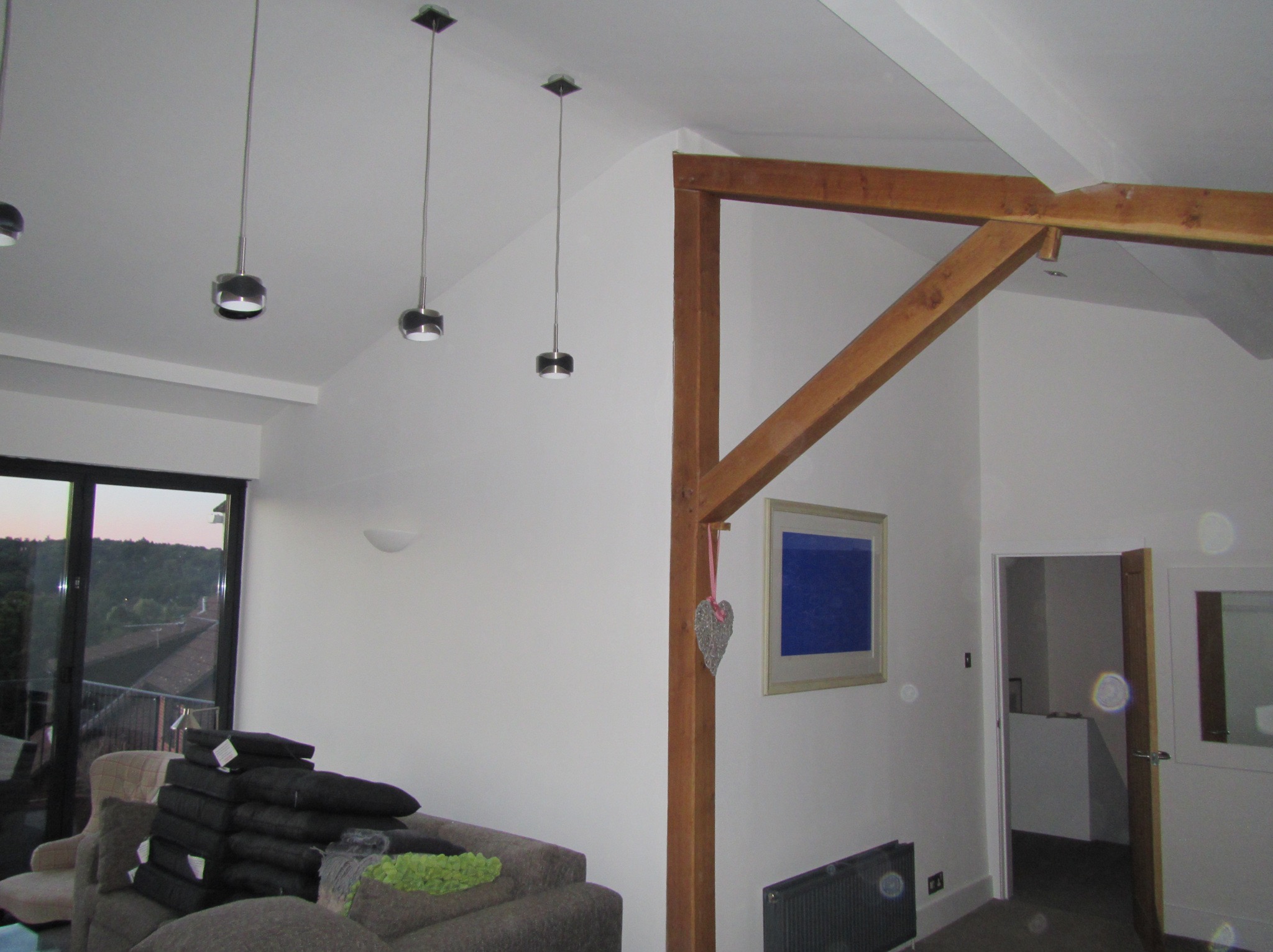 Wnętrze pokoju z drewnianą konstrukcją stropu, trzema wiszącymi lampami z metalowymi kloszami, stosem poduszek na kanapie i widokiem na zewnątrz przez duże okno.
