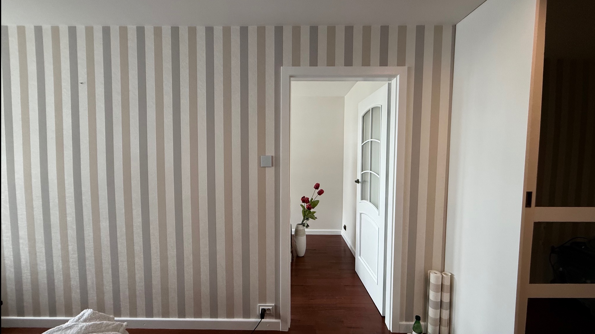 Pomalowany pokój w paski, z otwartymi drzwiami i widokiem na wazon z kwiatami. Widoczny fragment szafy wnękowej i podłoga z ciemnego drewna.