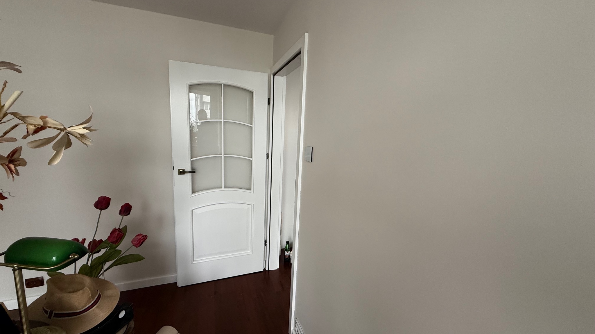 Jasnoszara ściana po malowaniu z białą, częściowo otwartą, przeszkloną  drzwią w tle. Na pierwszym planie kwiaty i lampka.