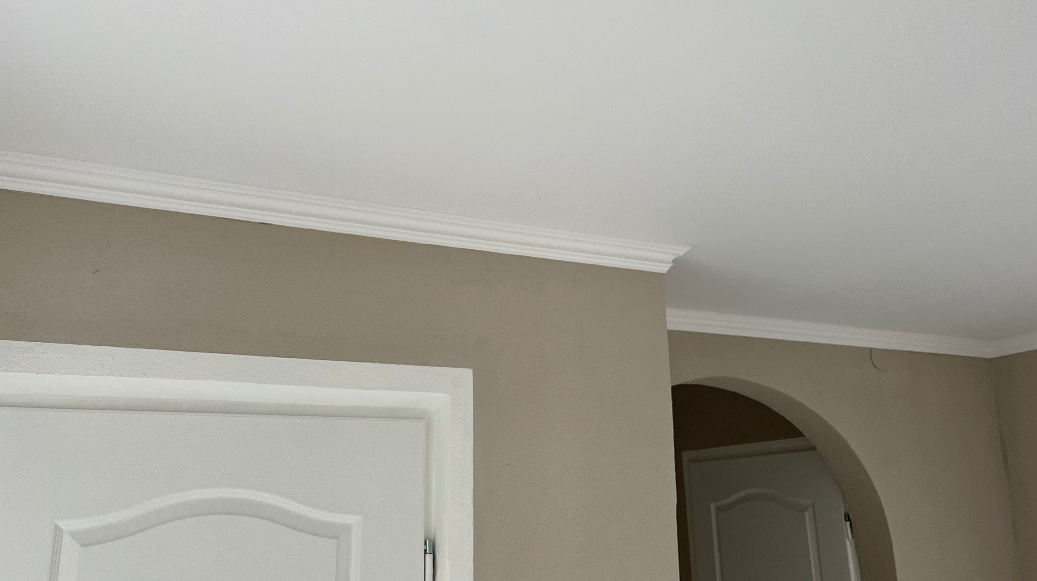 Ściana w odcieniach beżu z białym sufitem i sztukaterią. Widoczny fragment białych drzwi i łukowatego przejścia do innego pomieszczenia. Neutralne, świeże wykończenie wnętrza.