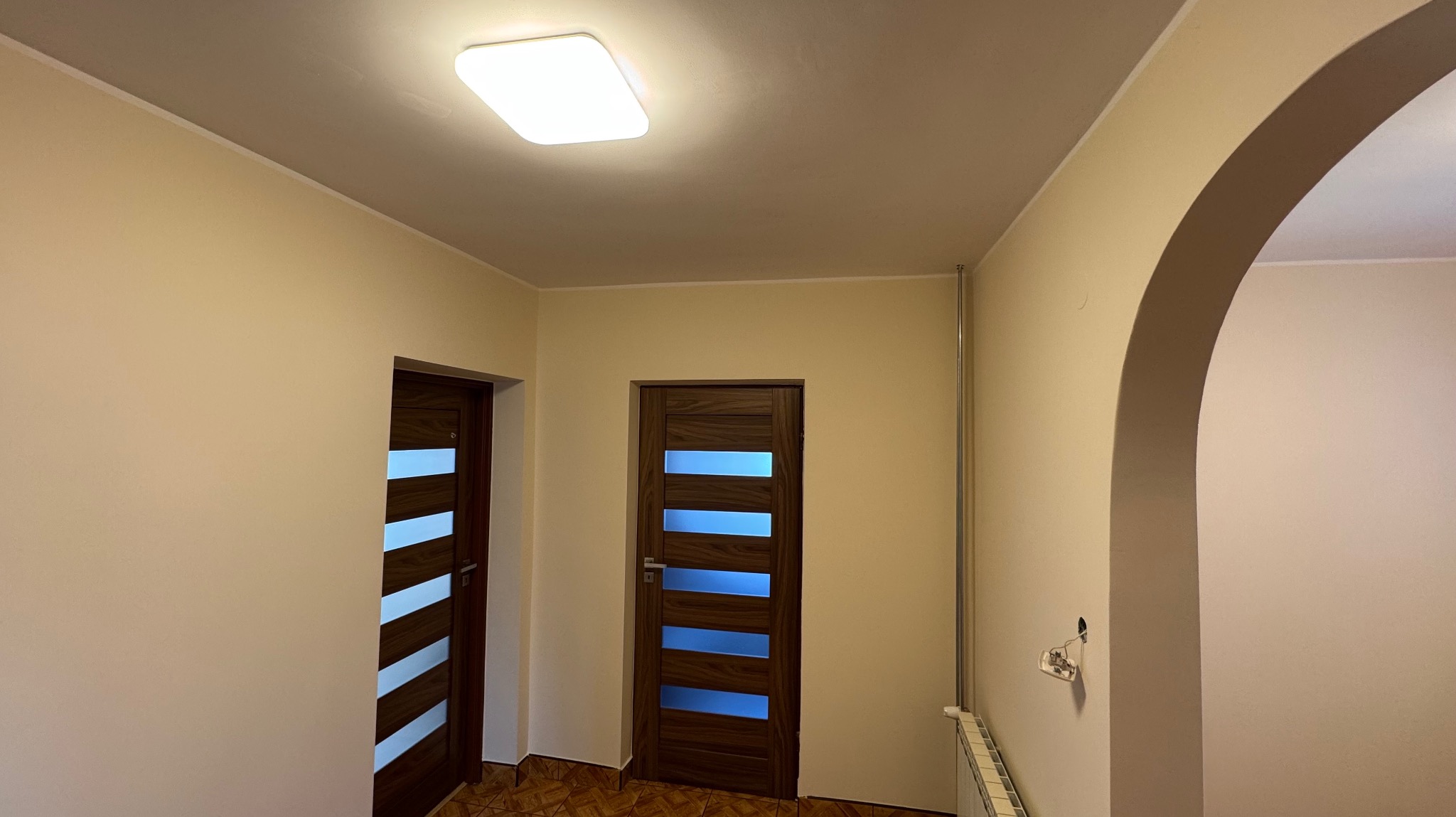 Jasne ściany po malowaniu w korytarzu z dwiema parami drzwi z przeszkleniami i łukiem. Widoczny grzejnik i gniazdko elektryczne.