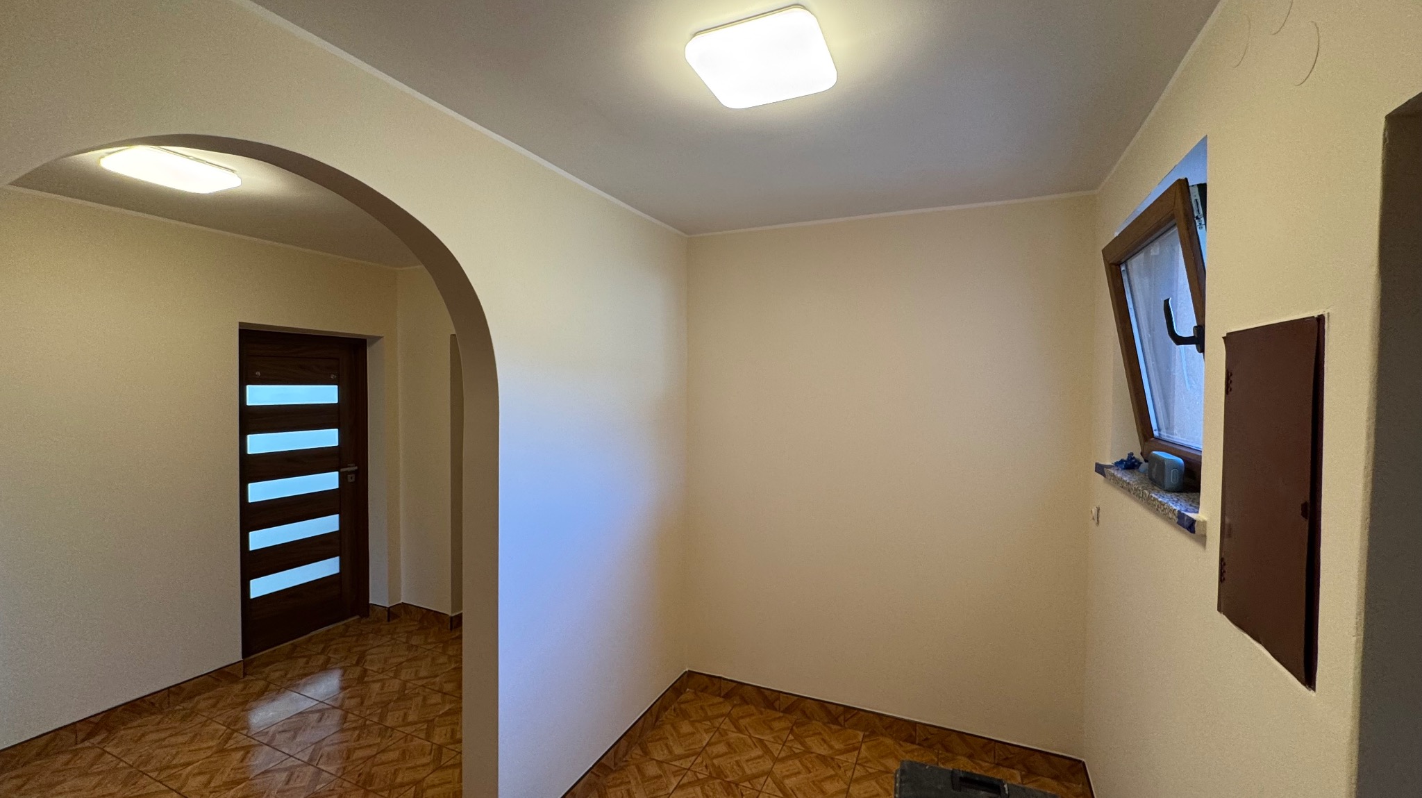 Jasne wnętrze po malowaniu: kremowe ściany, brązowe płytki podłogowe, drewniane drzwi z przeszkleniami i otwarte okno. Widoczny łuk wejściowy.