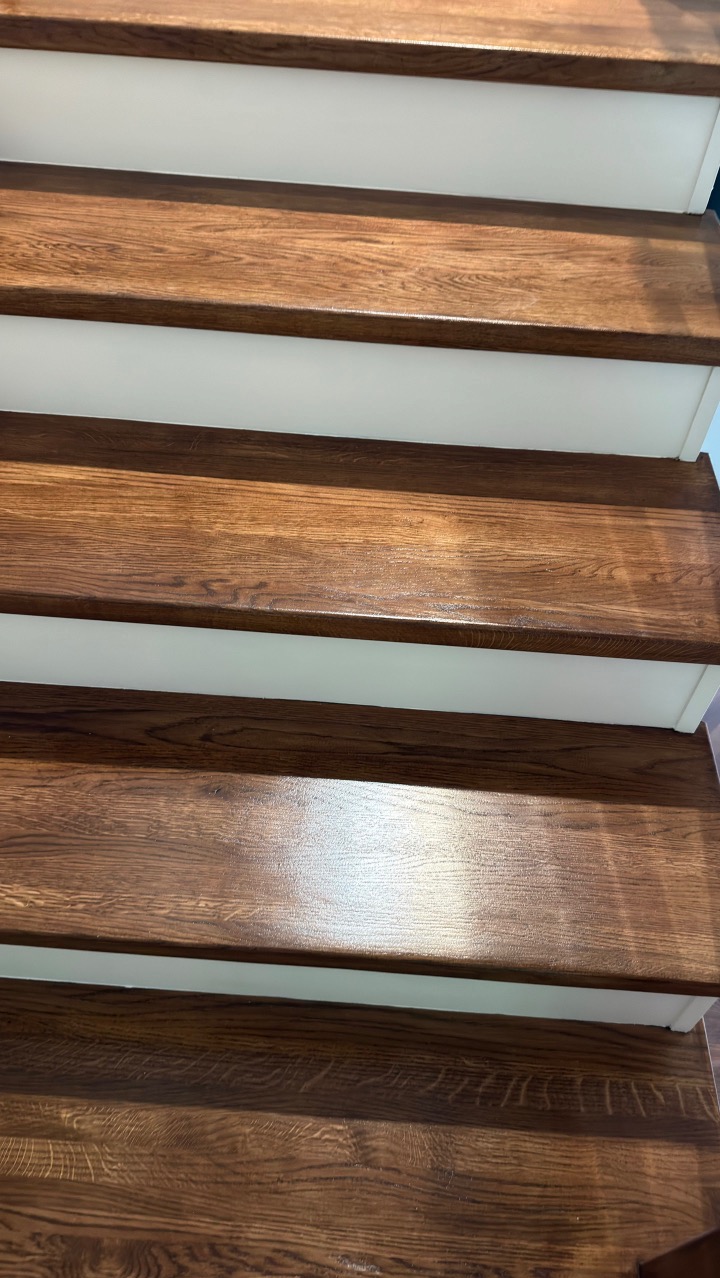 Błyszczące, drewniane schody z białymi podstopnicami, ukazane z bliska pod kątem. Widoczne detale słojów drewna i odbicia światła na powierzchni stopni.