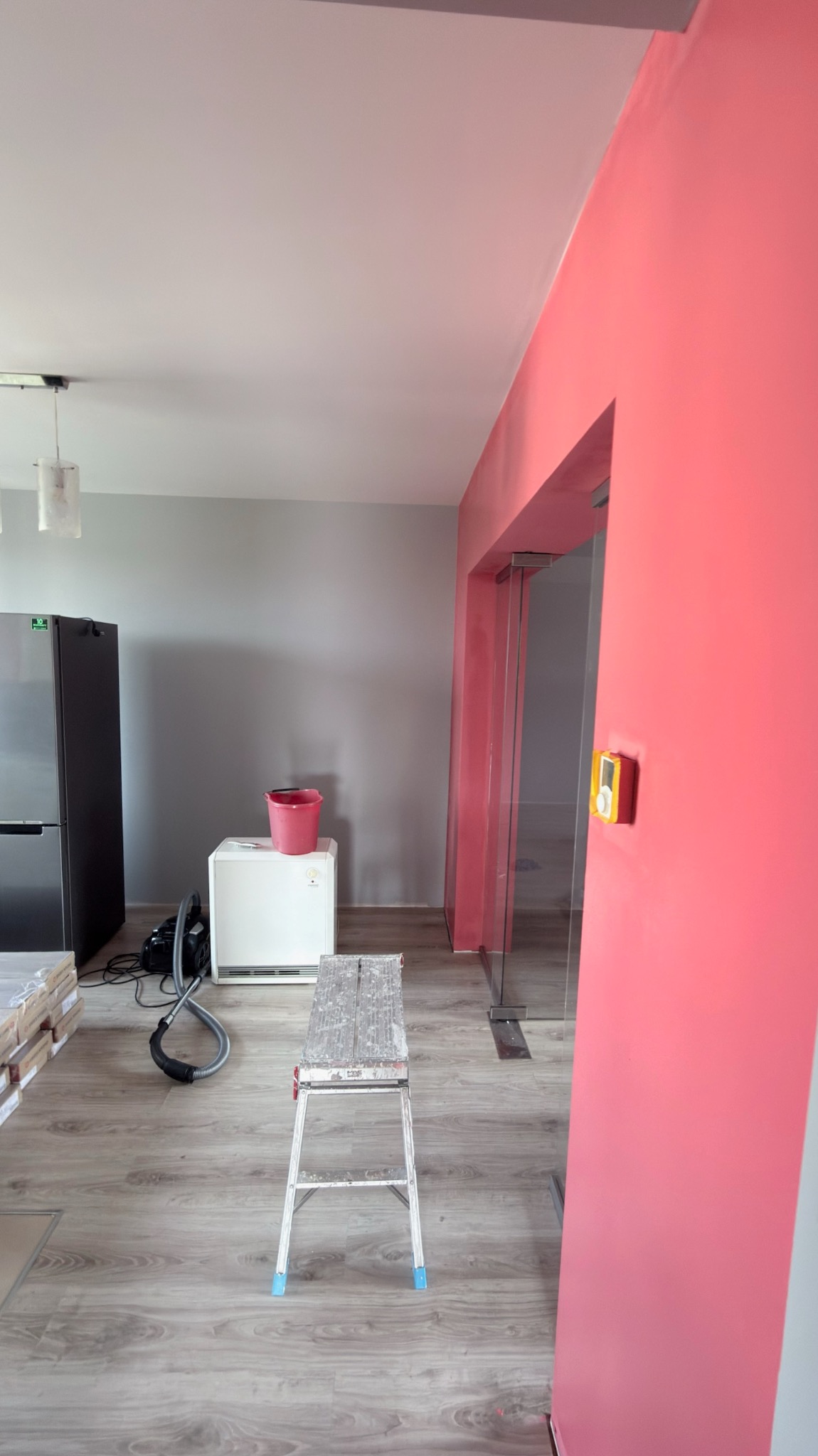 Pokój po malowaniu: ściana w odcieniach różu, obok szara ściana. Widoczna drabina, wiadro i odkurzacz. Podłoga drewniana. Prace wykończeniowe w trakcie.