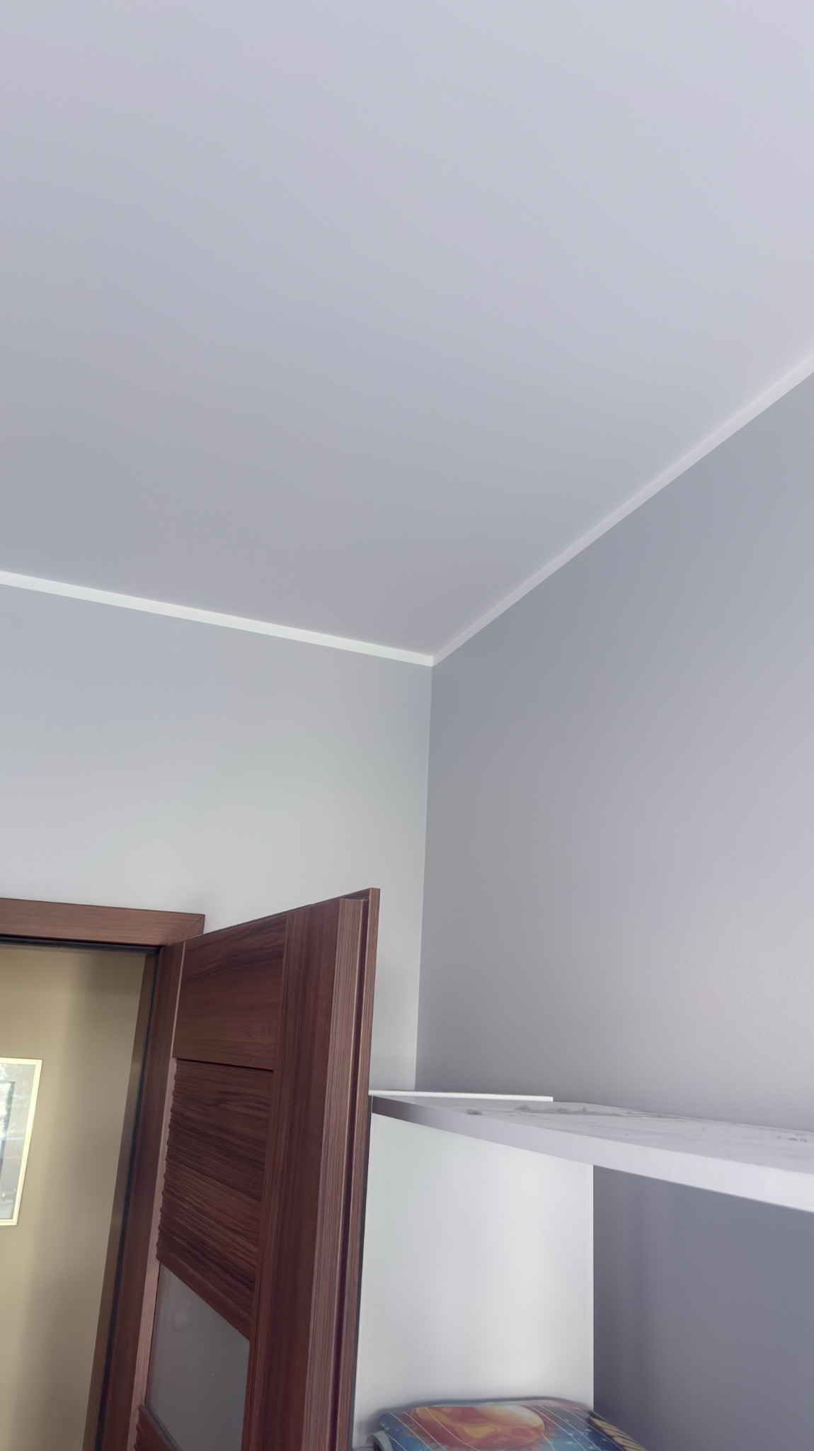 Ściana i sufit w odcieniach szarości po malowaniu, widoczna listwa przypodłogowa i fragment drewnianych drzwi. Ujęcie z dołu, podkreśla gładką powierzchnię.