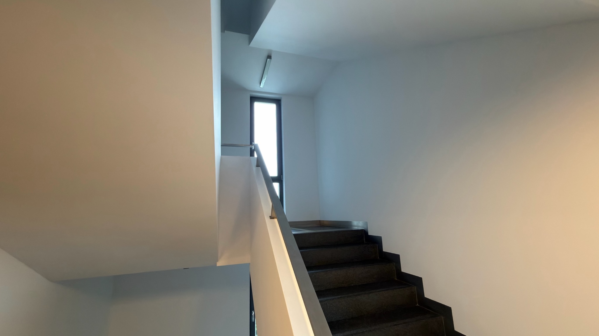 Klatka schodowa po malowaniu: białe ściany, czarne schody, metalowa poręcz. Widok z dołu, światło dzienne z okna na końcu biegu schodów.