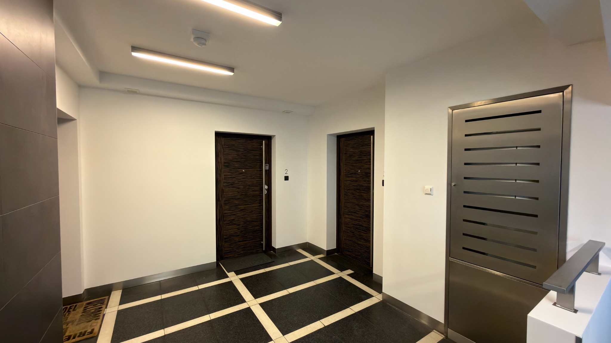 Wnętrze korytarza po malowaniu: białe ściany, ciemne drzwi z numerem '2', czarno-biała podłoga. Widoczne oświetlenie sufitowe i metalowa szafka.
