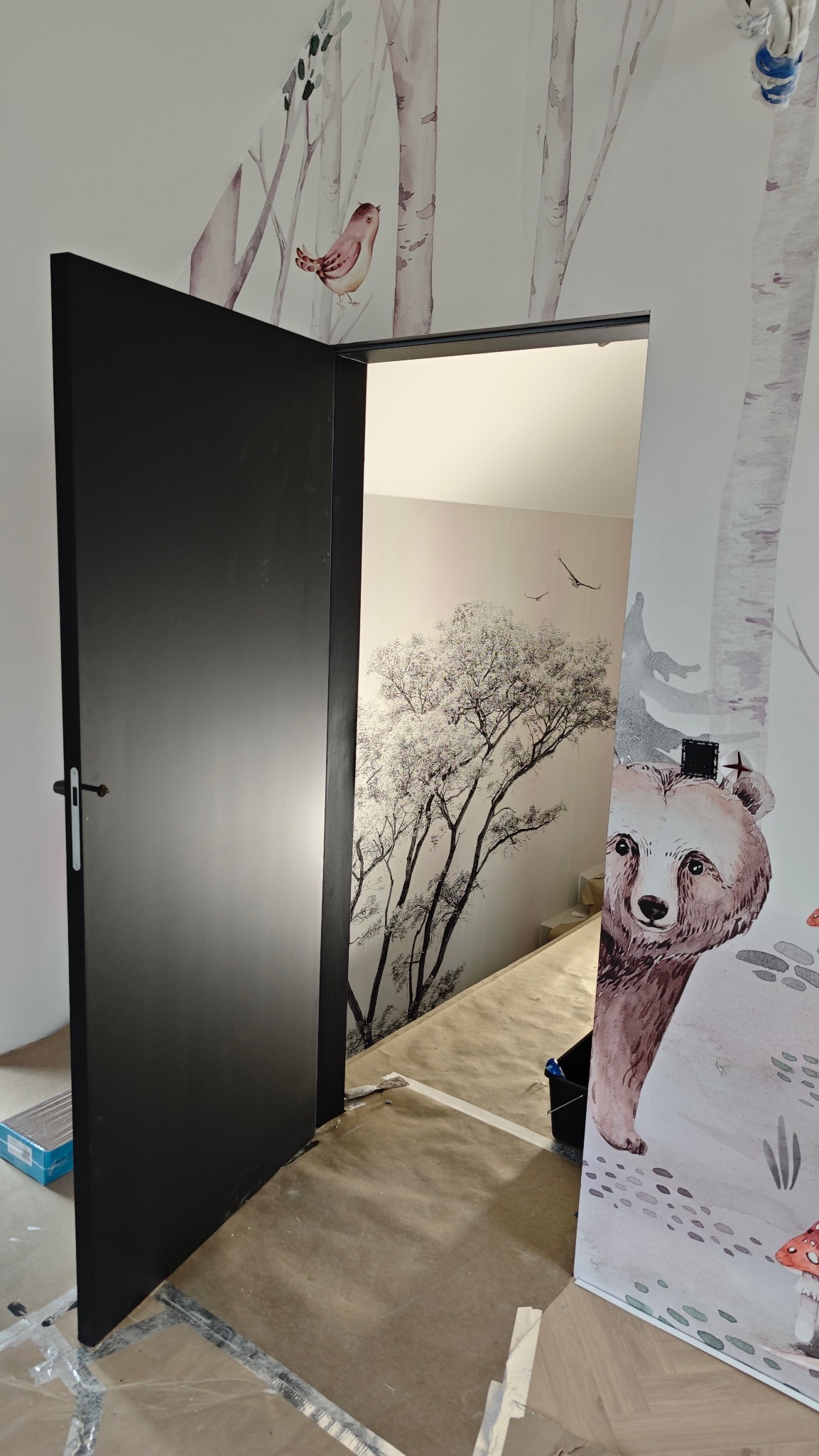 Tapetowanie pokoju dziecięcego: fototapeta z motywem lasu i niedźwiedzia, czarne drzwi. Widoczne zabezpieczenia podłogi. Realizacja w trakcie.