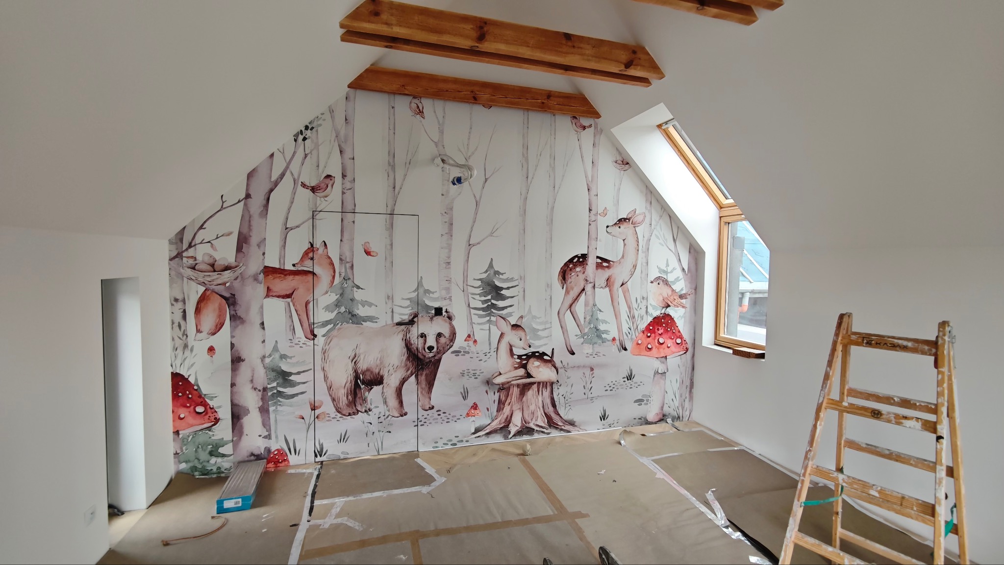 Pokój na poddaszu w Katowicach po tapetowaniu. Na ścianach tapeta w leśne zwierzęta, drewniane belki na suficie, okno dachowe i drabina malarska.
