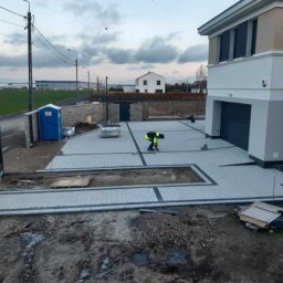 julbruk - Pracownik w odblaskowej kamizelce układa szarą kostkę brukową z ciemnymi obrzeżami na podjeździe przed nowoczesnym domem z garażem, widoczna przenośna toaleta i przygotowany teren.