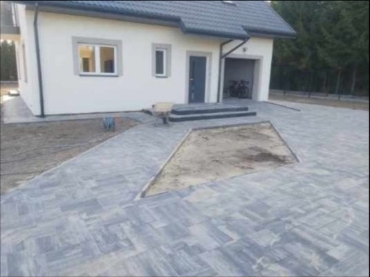 Szara kostka brukowa ułożona przed nowoczesnym domem z garażem, fragment terenu przygotowany pod dalsze prace, widoczny wózek budowlany.