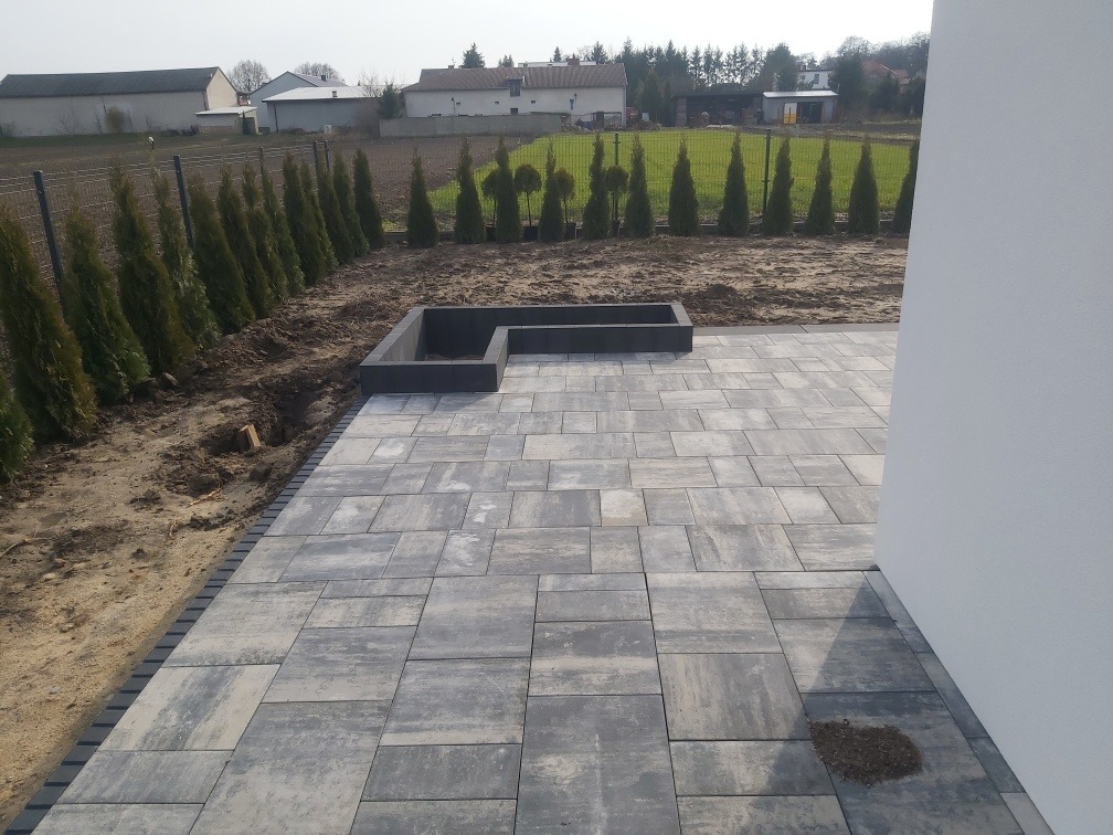 Ułożona szara kostka brukowa na tarasie z obrzeżami, z podniesioną rabatą w kształcie litery L, rząd tuj w tle, widok z boku budynku.