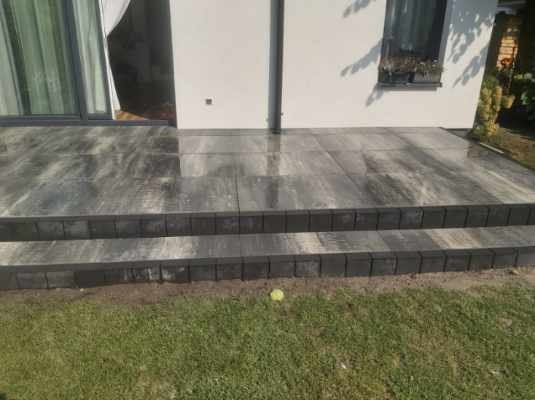 Wykończony taras z kostki brukowej w odcieniach szarości, z dwoma stopniami, przylegający do białego budynku z dużymi oknami, widok z poziomu trawnika.
