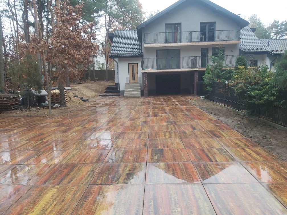 Ułożona, mokra nawierzchnia z wielkoformatowych płyt imitujących drewno, prowadząca do domu z garażem i balkonami, widok z perspektywy niskiego kąta.