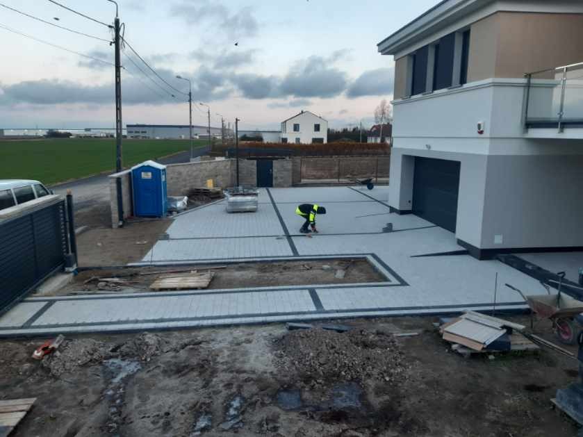 Pracownik w odblaskowej kamizelce układa szarą kostkę brukową z ciemnymi obrzeżami na podjeździe przed nowoczesnym domem z garażem, widoczna przenośna toaleta i przygotowany teren.