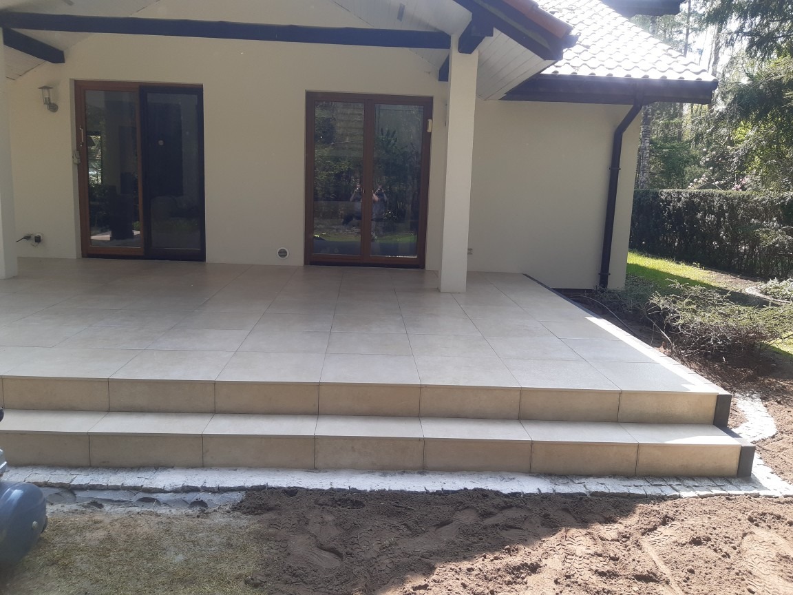 Beżowa, ceramiczna nawierzchnia tarasu z dwoma stopniami, przylegająca do jasnego budynku z brązowymi drzwiami przesuwnymi; widoczne obrzeże z jasnej kostki i fragment przygotowanego podłoża...