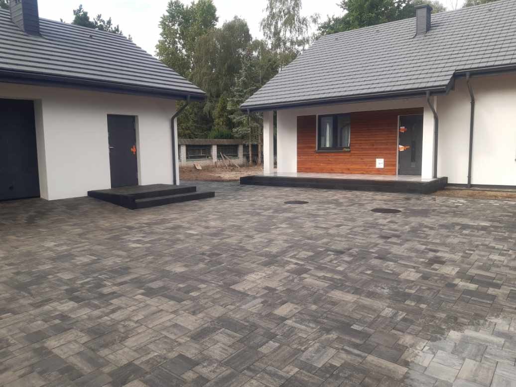 Szeroki podjazd z szarej kostki brukowej ułożonej we wzór jodełki, prowadzący do dwóch budynków z białymi ścianami i ciemnymi dachami. Widoczne włazy kanalizacyjne w kostce. Jeden budynek...