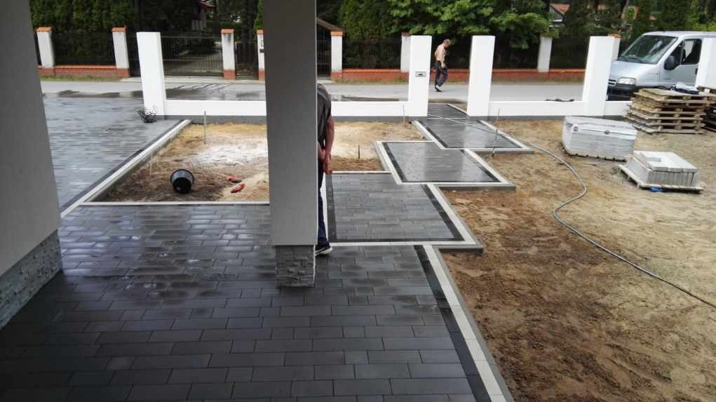 Częściowo ułożona, ciemnoszara kostka brukowa na podjeździe domu z elementami dekoracyjnymi, widoczne palety z kostką i przygotowane podłoże pod dalszą część nawierzchni.