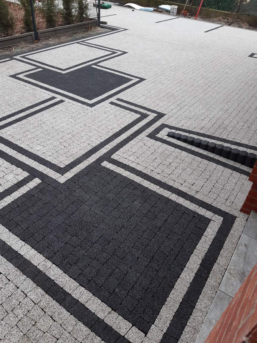 Ułożona kostka brukowa w odcieniach szarości i czerni, tworząca geometryczny wzór z kwadratami i liniami, widoczny drenaż liniowy z okrągłych elementów, ogrodzenie panelowe w tle.